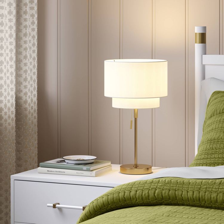 A lamp with a white colored shade and gold colored base is placed on a white bedside table next to a bed with a green colored quilt.