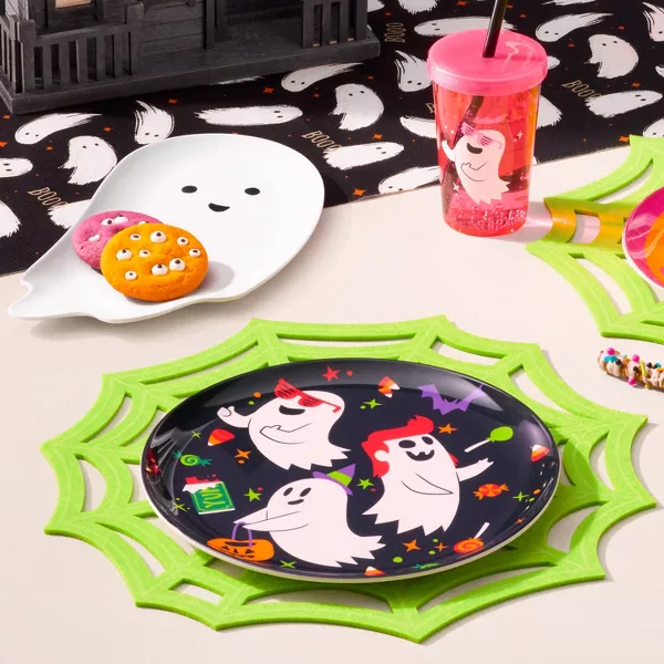 This party decor is kid-friendly & bright. A smiling ghost, pumpkin, & black round plate with 3 ghosts' trick or treating sit on a table holding cookies with eye candies. Green spiderweb placemats are used along with a black table runner with ghosts.