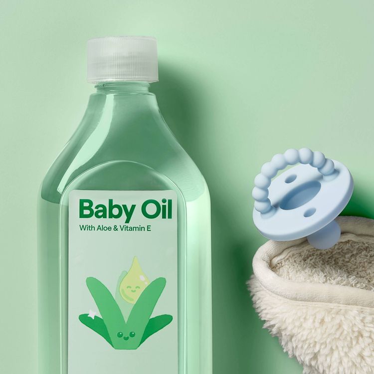 A clear plastic bottle labeled "Baby Oil with Aloe & Vitamin E" next to a light blue pacifier and a fluffy beige cloth on a light green background.