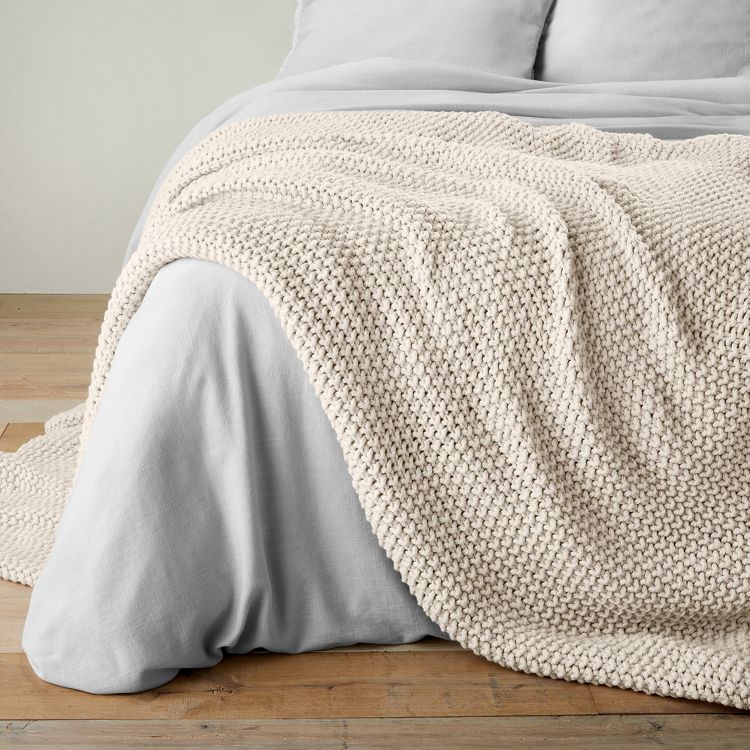 A natural-colored chunky knit bed blanket draped over a bed with white sheets, placed on a wooden floor.