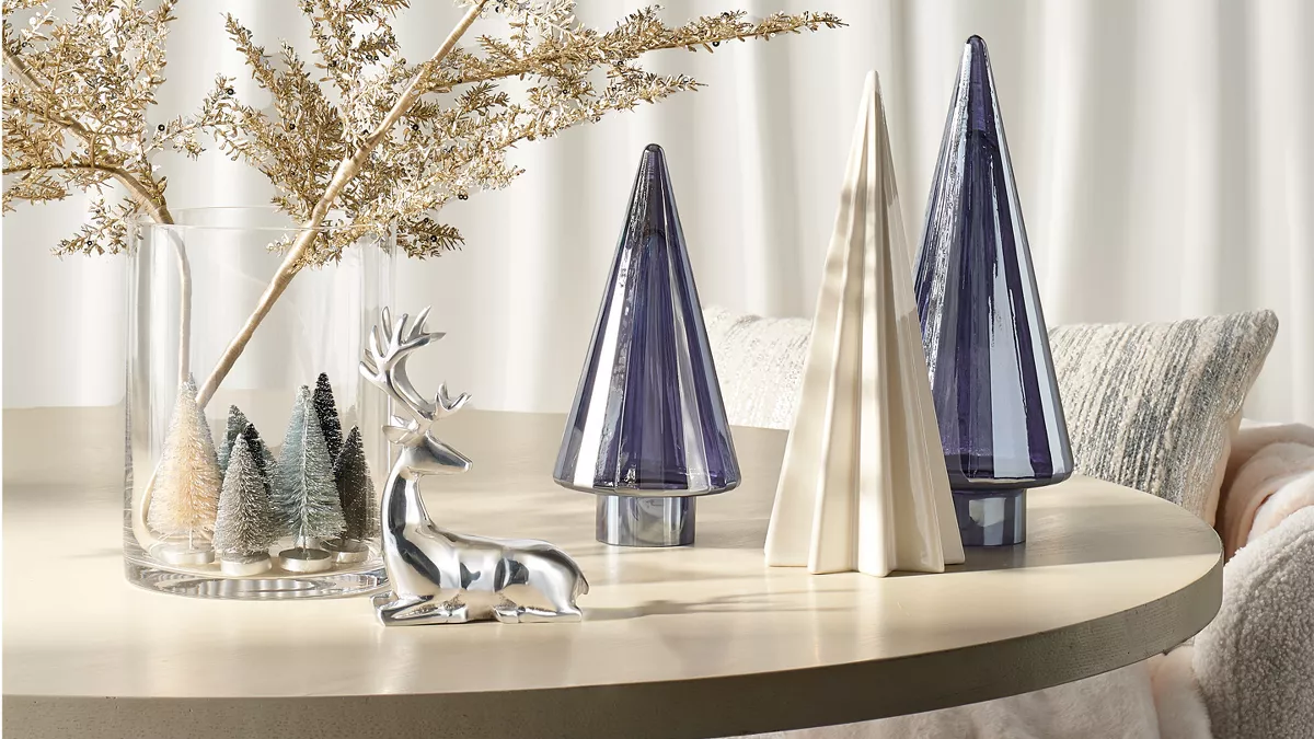 1 white ceramic & 2 metallic blue Christmas trees sit atop a table. A silver reindeer figurine sits in front of a large glass vase. Inside the vase, mini white & green Christmas trees line the bottom & large gold branches expand out. 