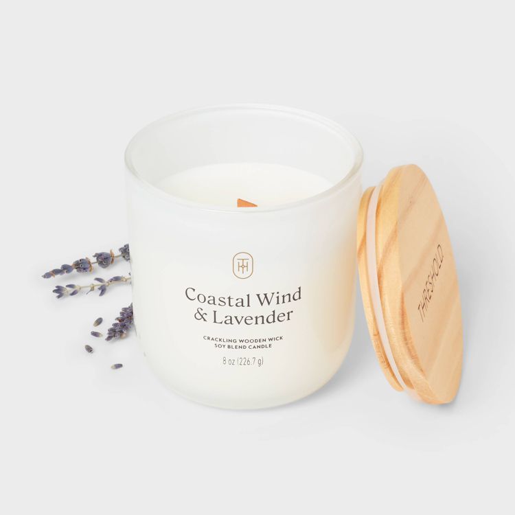 A white colored candle in a glass jar labeled "Coastal Wind & Lavender" with a wooden lid and sprigs of lavender beside it.