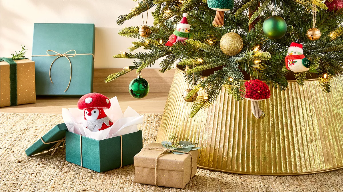 A decorated Christmas tree with green and gold ornaments and cute figures stands on a gold-colored tree stand. Three wrapped gifts, one open with a mushroom-shaped toy inside, are placed on a tan woven rug next to the tree.