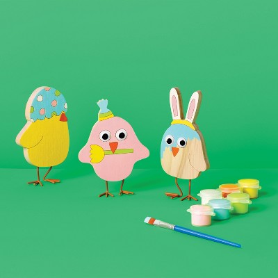 Three painted wooden bird figures with googly eyes and a paintbrush beside small pots of various colored paints on a green background.