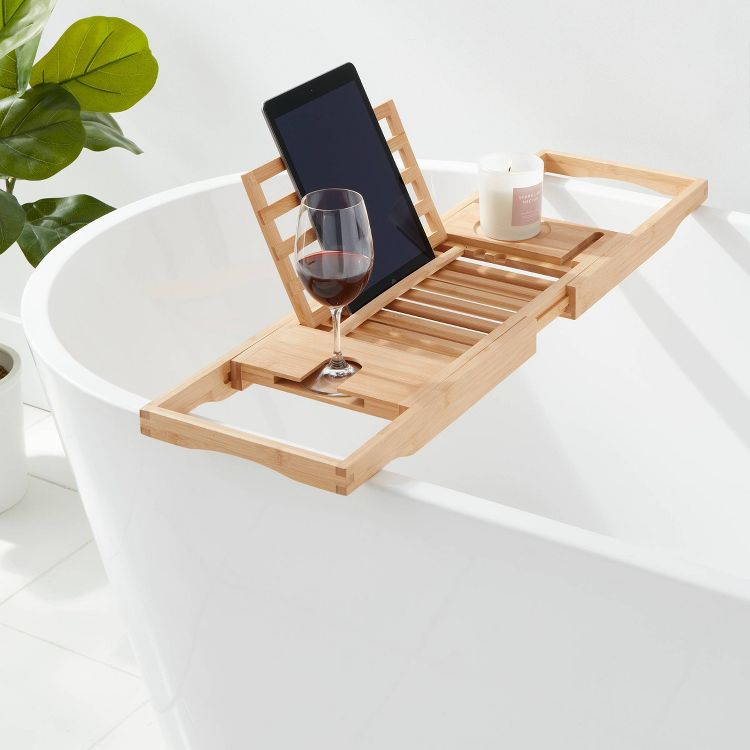 A wooden bath caddy rests across a white bathtub, holding a tablet, a glass of red wine, and a lit candle.
