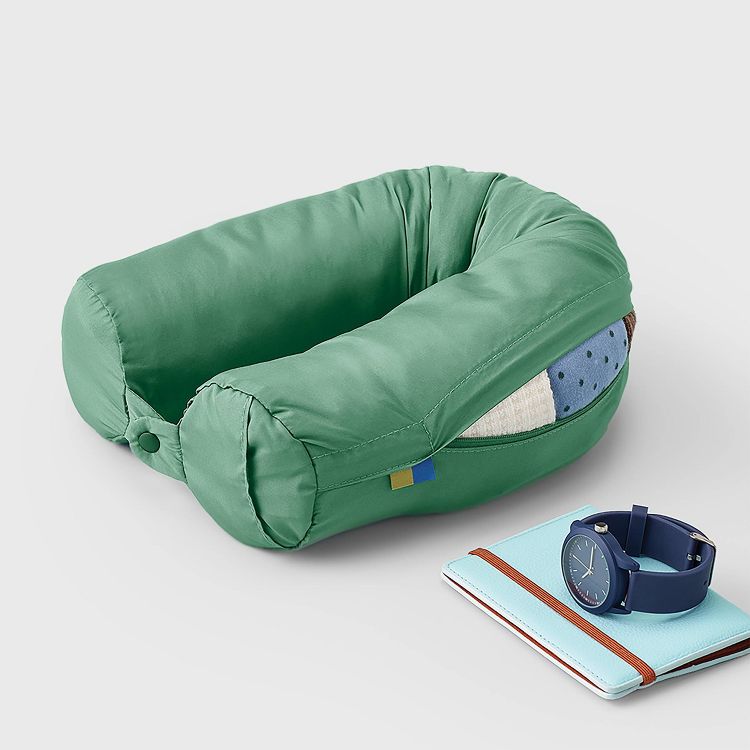 A green colored travel neck pillow with a button closure, placed next to a blue watch and a light blue notebook.