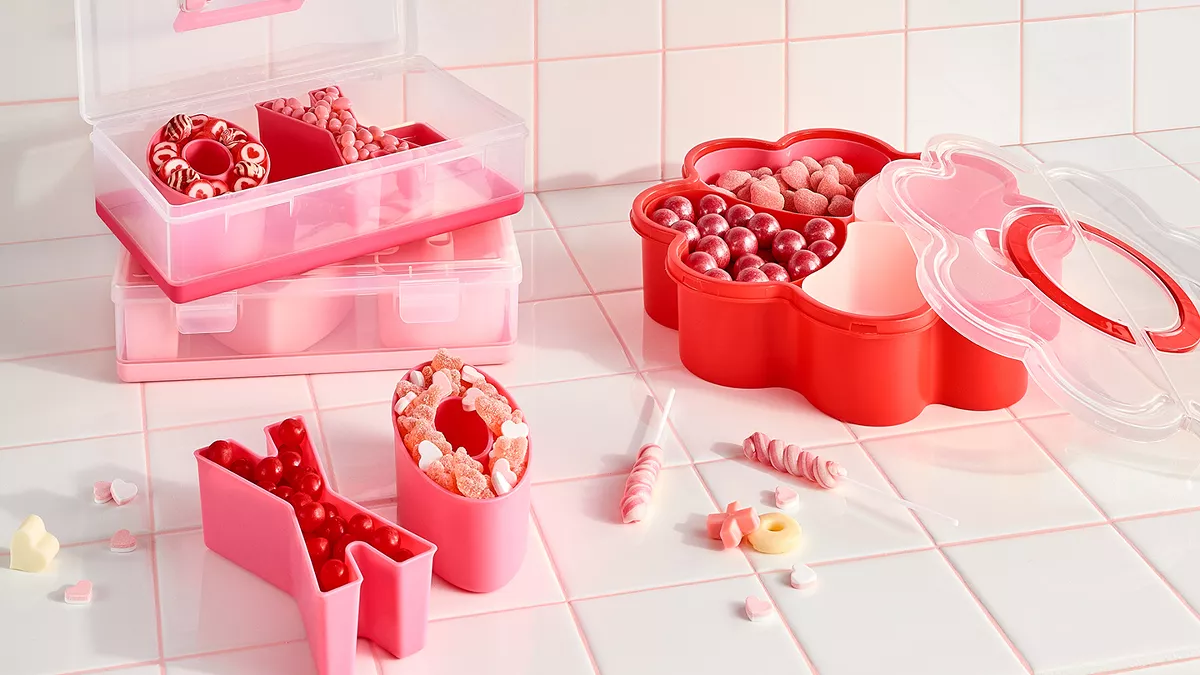 Red & pink candies fill up V-Day shaped bins. There is a pair of X & O bins, a larger bin made up of 4 separate hearts & 2 plastic rectangle bins with red & pink lids. They all sit on a white tile counter with some candies spilled over. 