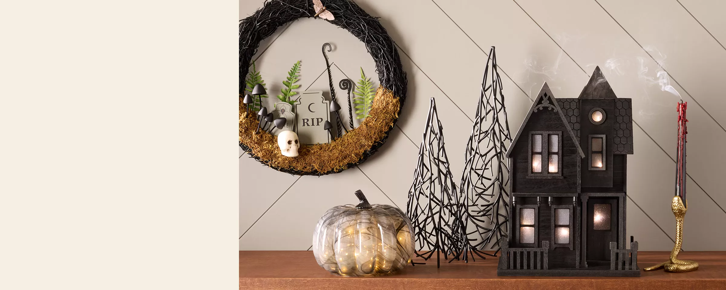 A black 2-story haunted house with lights sits on a table between a gold snake candle holder, 2 black metal trees & a clear glass pumpkin with twinkling lights inside. On the wall, a black twig wreath hangs showing a graveyard scene. 