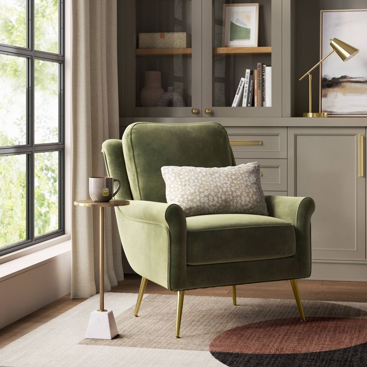 A small round table with a gold colored top and a white base, holding a mug, is placed beside a green colored armchair.