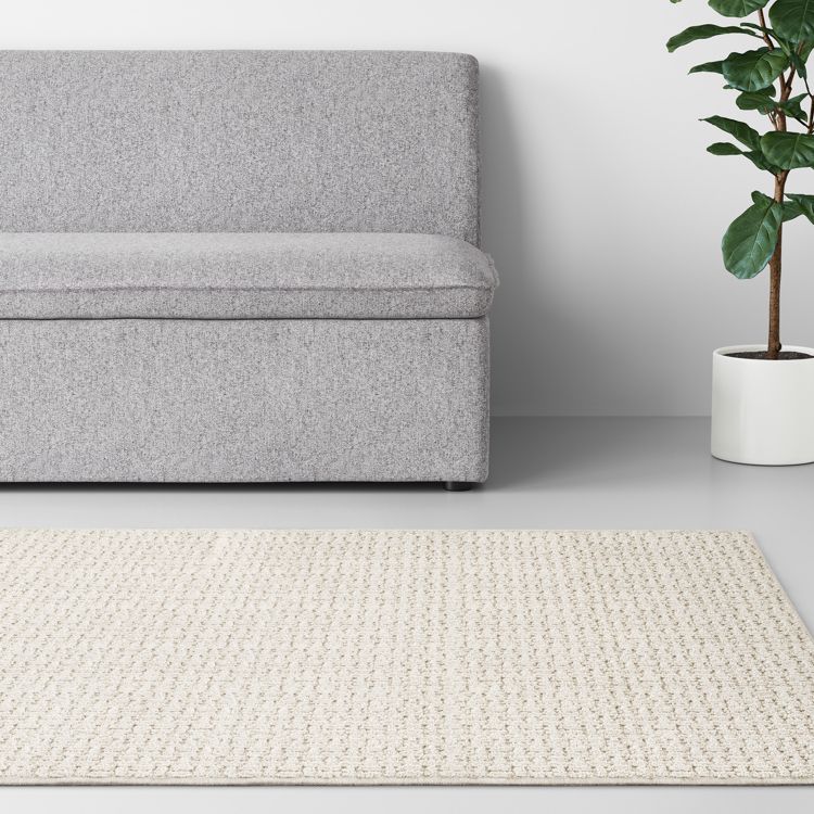 A beige colored textured rug placed on a light gray floor in front of a gray sofa and a potted plant.