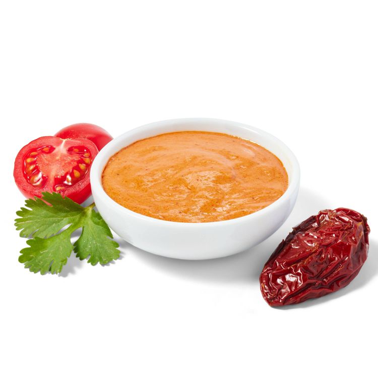 A bowl of orange colored sauce is surrounded by a halved tomato, a sprig of cilantro, and a dried red pepper.