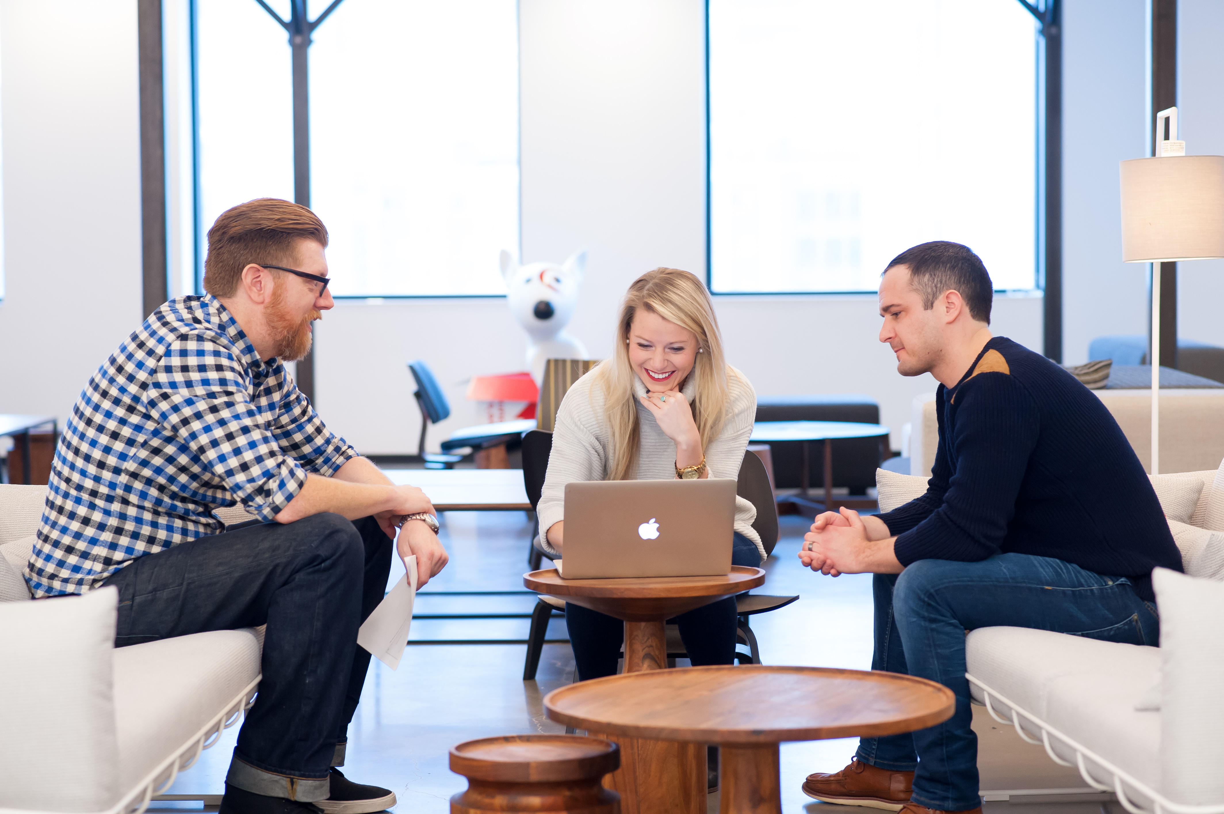 Interior shot of a meeting in a corporate Target office, with a man in a checked button down shirt sitting next to a woman looking at a laptop and smiling, and another man in a dark colored sweater sitting to her other side