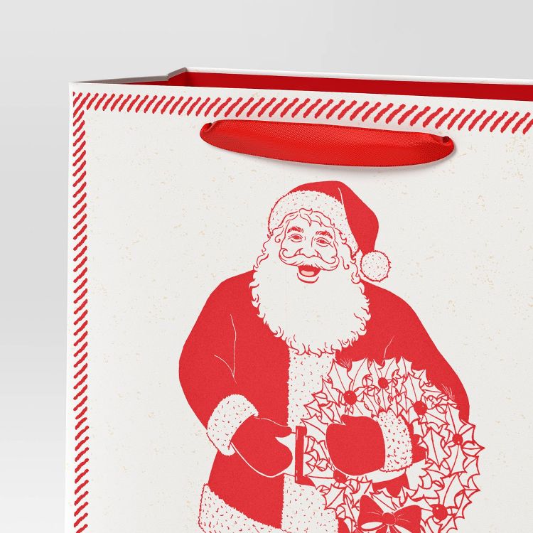 A white colored gift bag with a red colored illustration of Santa Claus holding a wreath, featuring a red handle and decorative border.