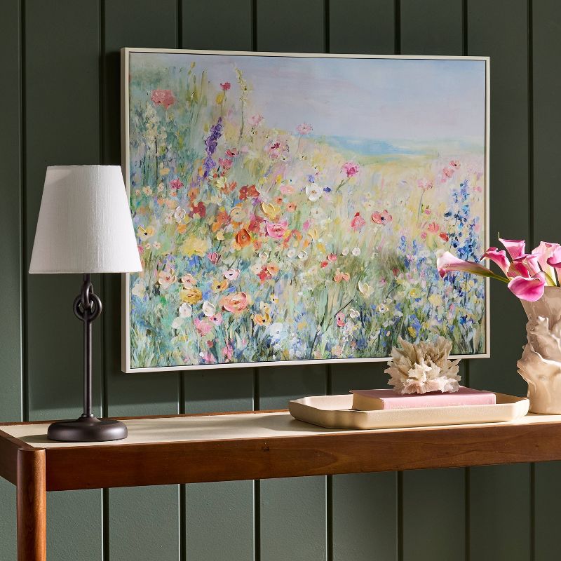 A colorful painting of a wildflower field is hung on a dark green paneled wall above a wooden console table with a lamp, a vase of pink flowers, and decorative items.