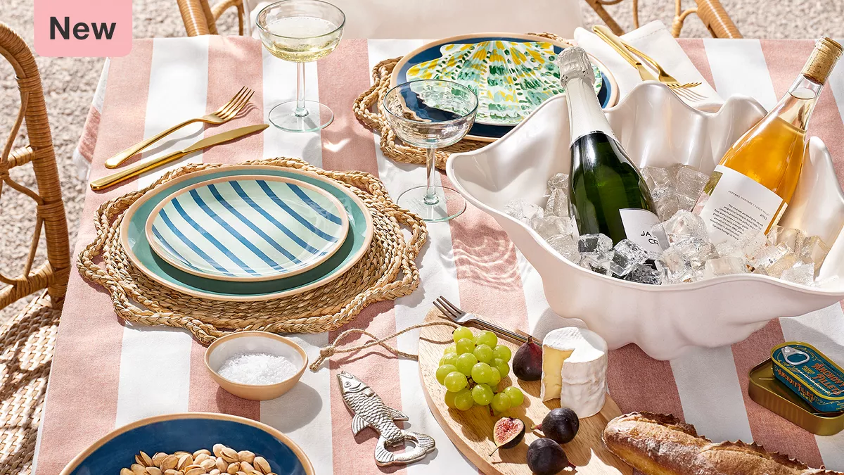 A cheerful outdoor table set with a striped tablecloth. There are dishes with blue stripes, nuts, grapes, figs, cheese, bread, and a shell-shaped cooler with drinks on ice. Gold-colored utensils and a tin can are also present.