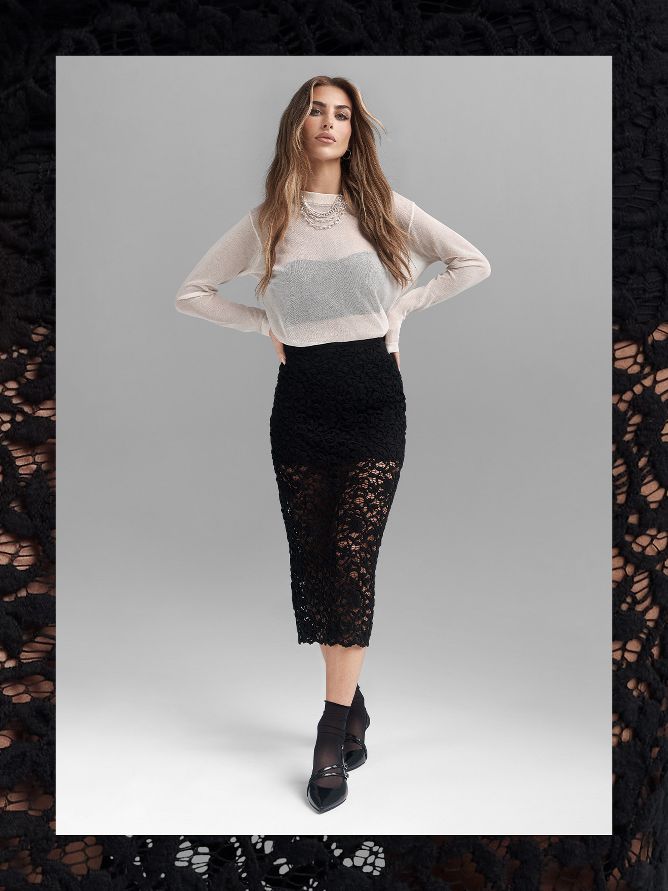 Paige Lorenze is rocking a black calf-length openwork skirt. On top, she wears a long-sleeve semi-transparent white shirt with a black cropped tank peeking out underneath. She finishes off the look with stacked necklaces and pointy black dress shoes.