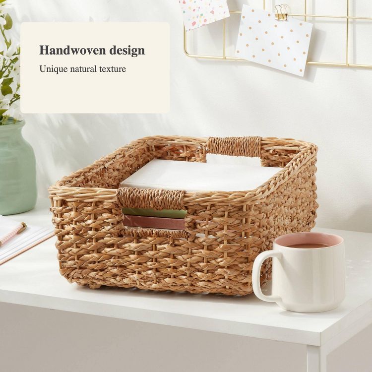 A natural colored handwoven basket with handles, placed on a white surface next to a white mug.