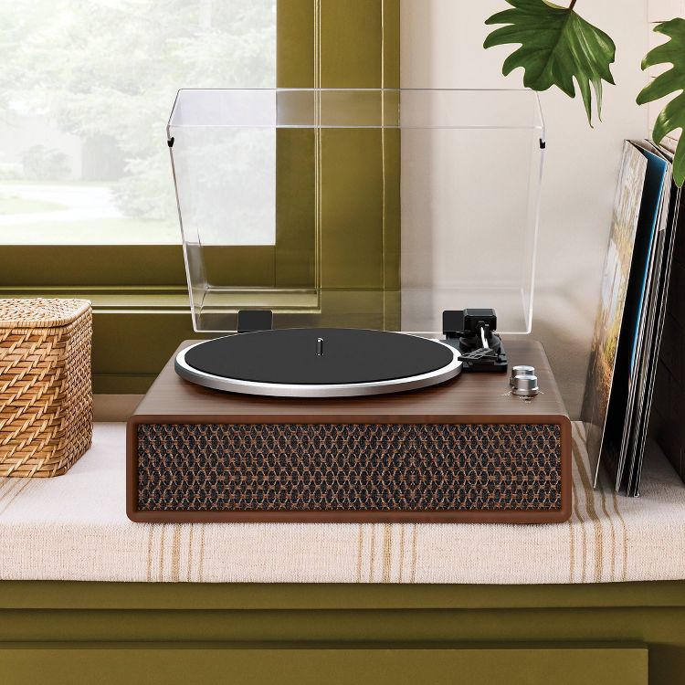 A brown colored turntable with a clear dust cover and a black platter, placed on a striped fabric surface near a window.