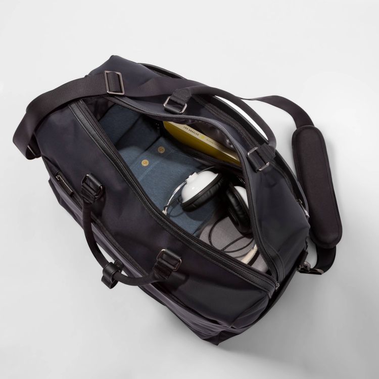A black colored duffel bag with an open top showing packed items including a pair of headphones, a book, and clothing.
