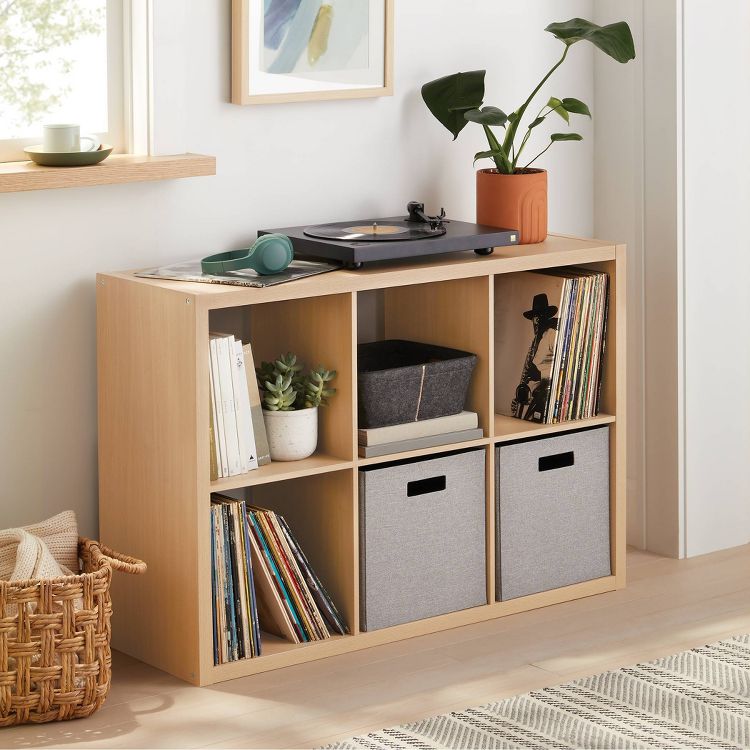 Natural wood cube organizer with six shelves, featuring books, plants, a record player on top, and two gray storage bins.