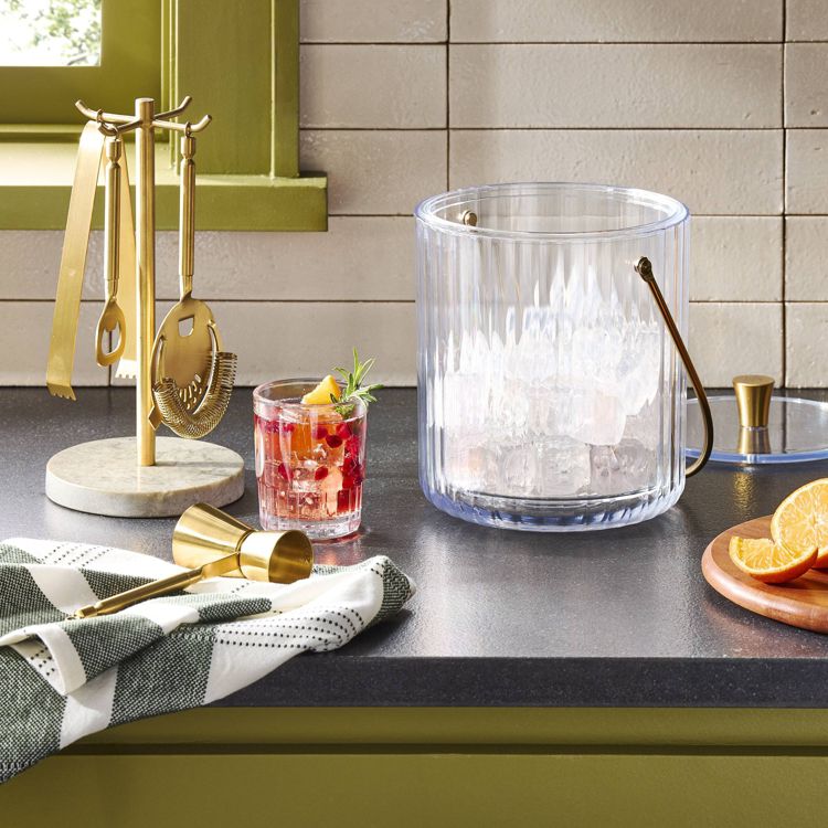 A clear ice bucket with a gold colored handle is placed on a countertop next to a set of gold colored bar tools on a stand, a cocktail glass with garnish, and a wooden board with sliced oranges.