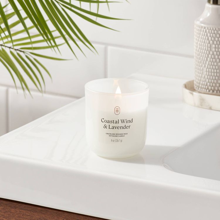 White candle labeled "Coastal Wind & Lavender" with a crackling wooden wick, placed on a white surface near a plant.