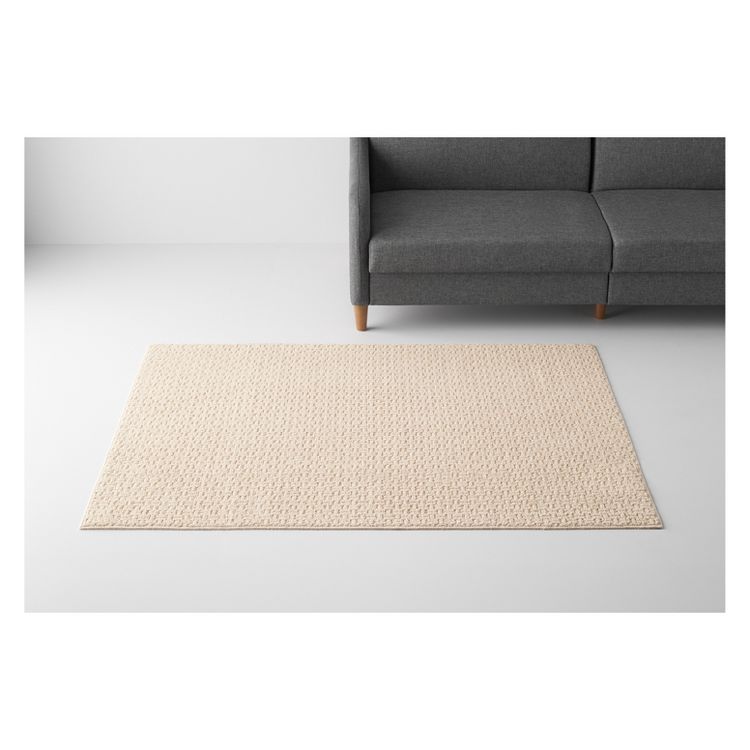A beige colored rectangular rug with a textured pattern is placed on a light gray floor next to a dark gray sofa.
