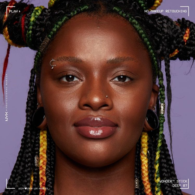 A person with glossy lip makeup and braided hair with green, yellow, and red colored sections, against a purple background, with text reading "NO MAKEUP RETOUCHING" and "WONDER STICK DEEP 07."