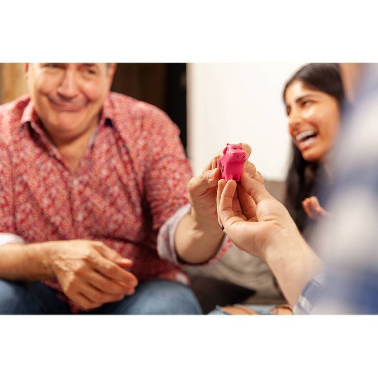 A person holds a small pink colored pig-shaped toy while others look on and laugh.