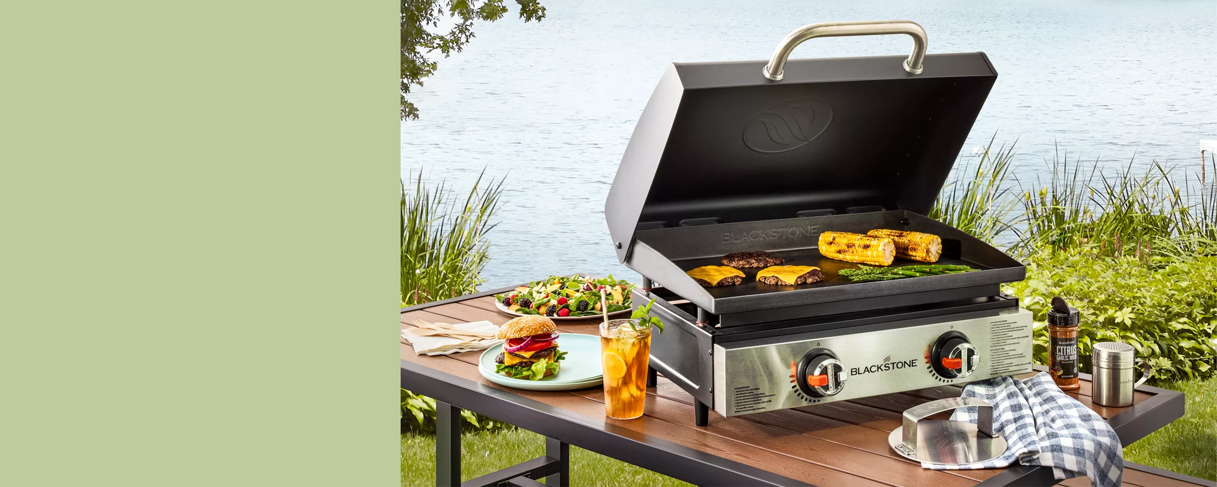 Outside near a serene pond, a portable Blackstone grill is on a patio table—filled with typical grilled meats & veggies. Grilling accessories & spices sit on the table, along with a refreshing iced tea, a berry-filled salad & assembled cheeseburger.