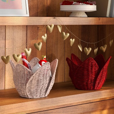 A pink colored woven basket with a tulip shape containing various items, placed next to a red colored basket on a wooden shelf, with a string of gold colored heart decorations above.