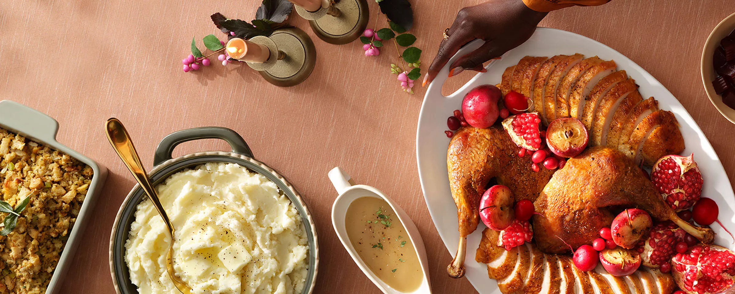 A dinner table with a platter of sliced turkey, garnished with pomegranate and small apples, is in the center. Surrounding it are mashed potatoes, green beans, a basket of bread, and drinks. The tablecloth is a soft brown color.