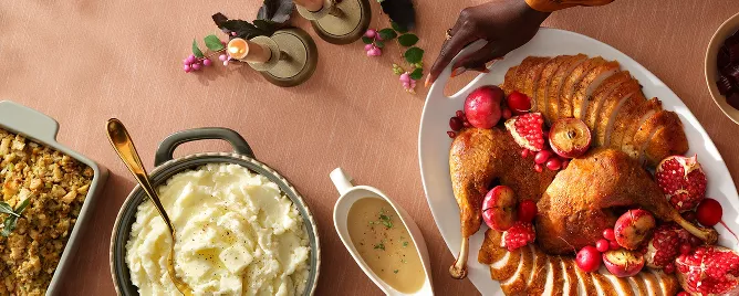 A dinner table with a platter of sliced turkey, garnished with pomegranate and small apples, is in the center. Surrounding it are mashed potatoes, green beans, a basket of bread, and drinks. The tablecloth is a soft brown color.