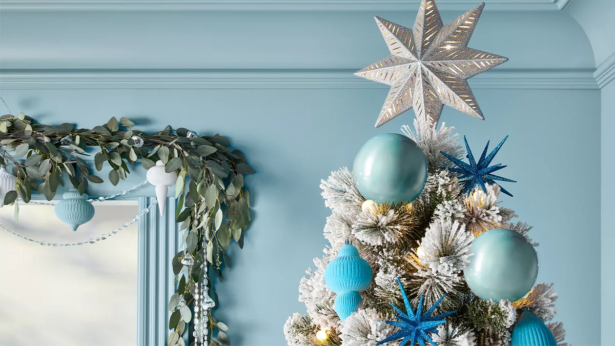 The image shows the top part of a Christmas tree with white-tipped branches. The tree is decorated with large blue and gold-colored ornaments, blue stars, and a bright star on top. Nearby, a garland with green leaves and white ornaments drapes over a window. The walls are light blue.