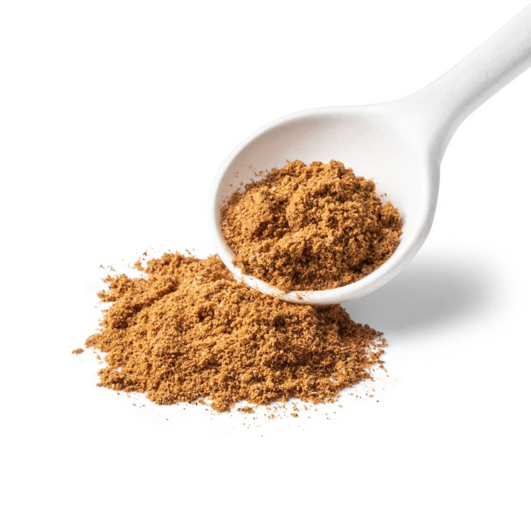 A white spoon filled with brown colored ground spice, with some spilled onto a white surface.