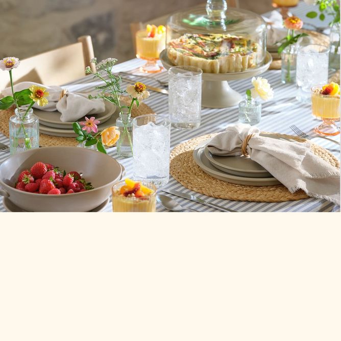 A dining table covered with a striped tablecloth in light blue and white, with place settings in natural tones. A quiche in a cakestand, a bowl of strawberries, and bud vases filled with spring flowers complete the scene.