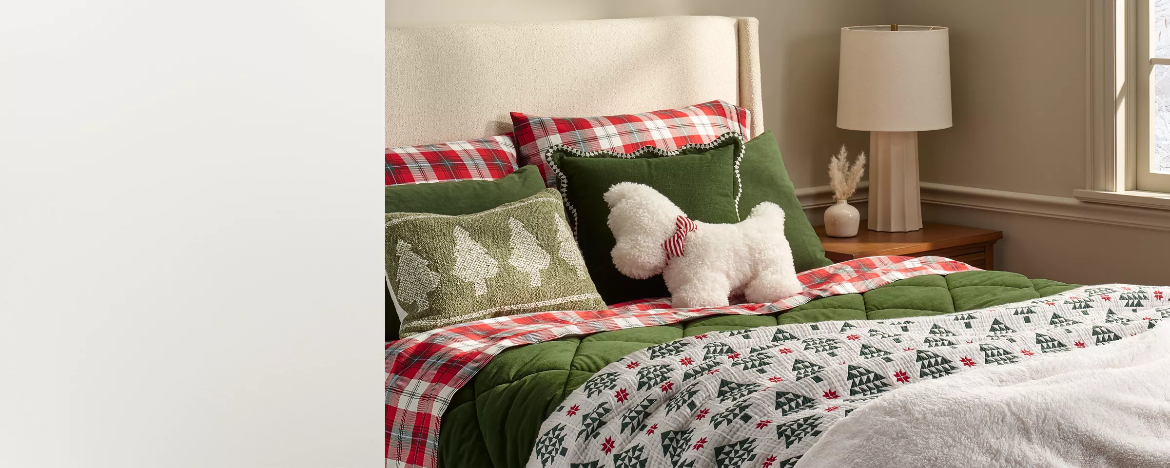 A bed is made with red & green plaid sheets & a green velvet comforter. A Christmas tree patterned quilt lays across the bottom of the bed along with a plush white blanket. A fuzzy white dog pillow with a bowtie rests at the top. It’s very festive & cozy.