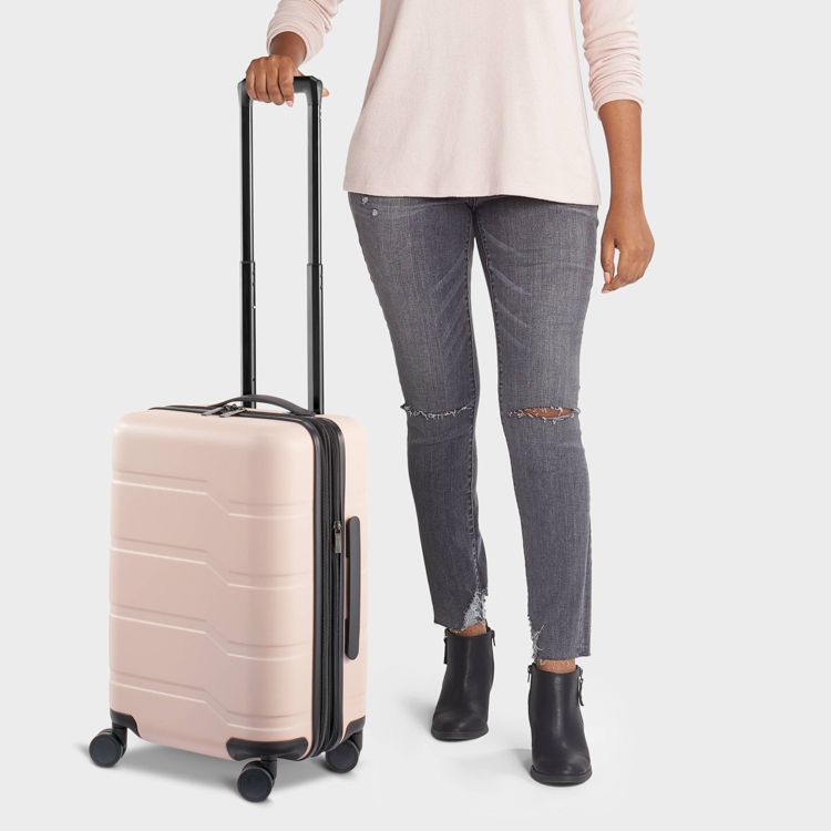 A light pink colored suitcase with a telescopic handle and four wheels being held by a person.