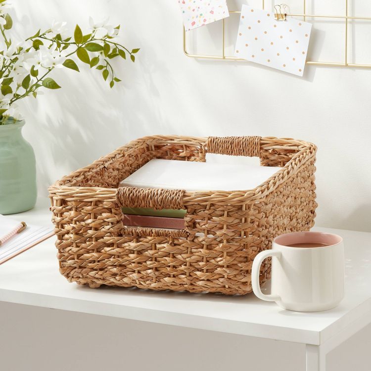 A woven basket with handles, containing papers, placed on a white desk next to a white mug.