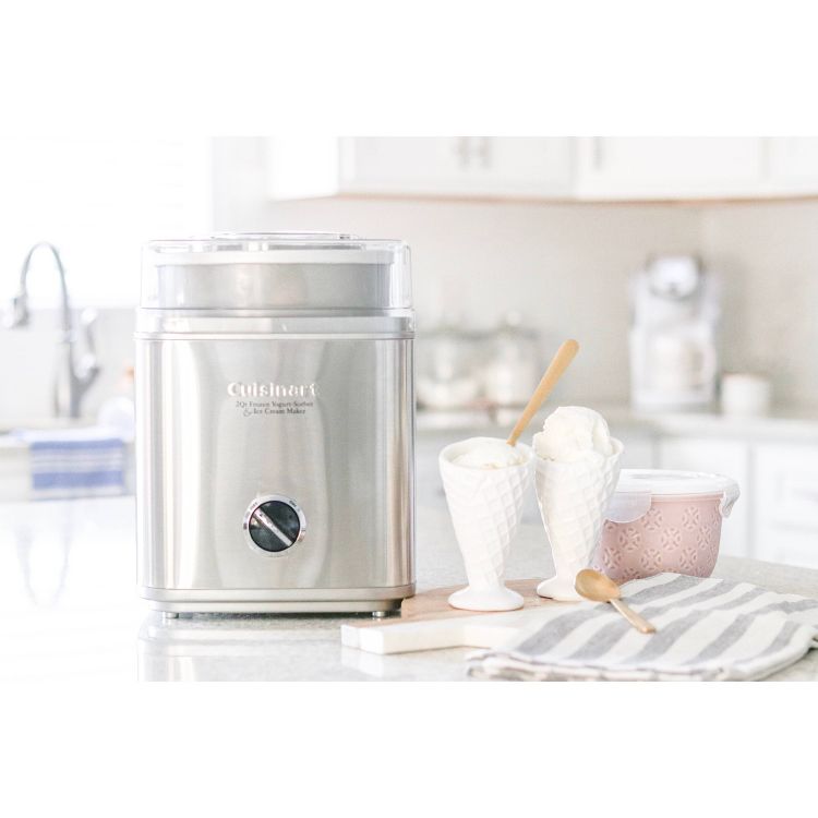 A stainless steel ice cream maker on a kitchen counter next to two white ice cream cups and a pink container.