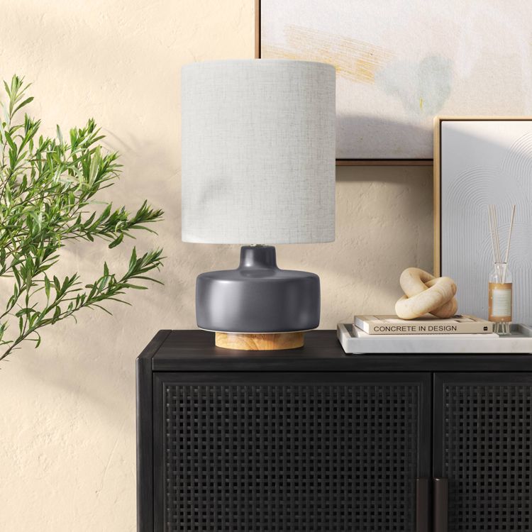 A table lamp with a light gray colored cylindrical shade and a dark gray colored base sits on a black cabinet.