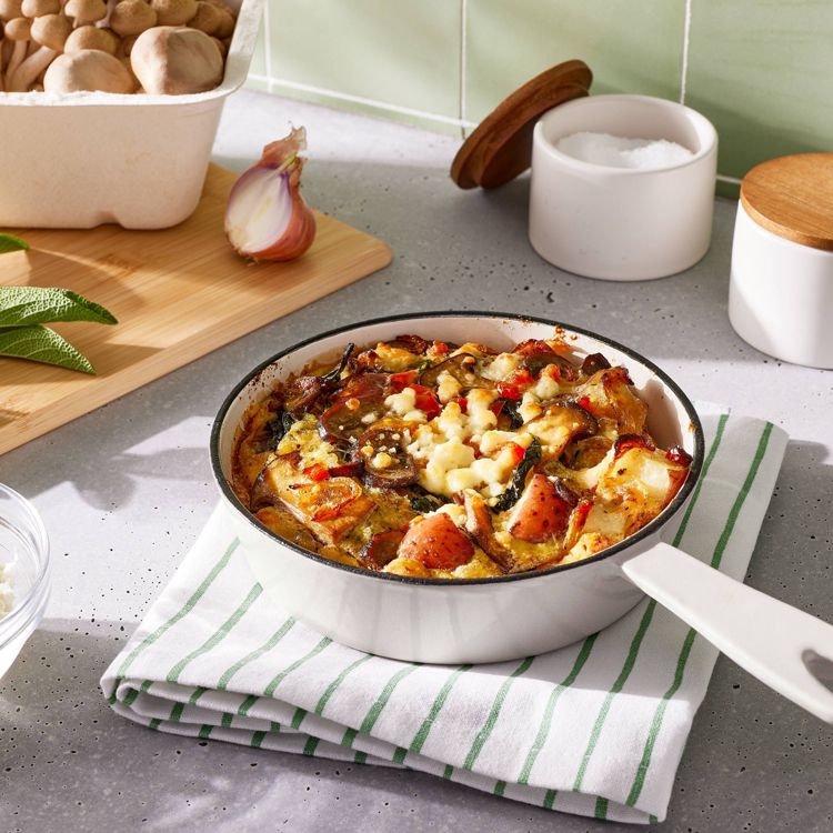 A white skillet filled with a baked dish containing vegetables and cheese, placed on a striped cloth on a kitchen counter.