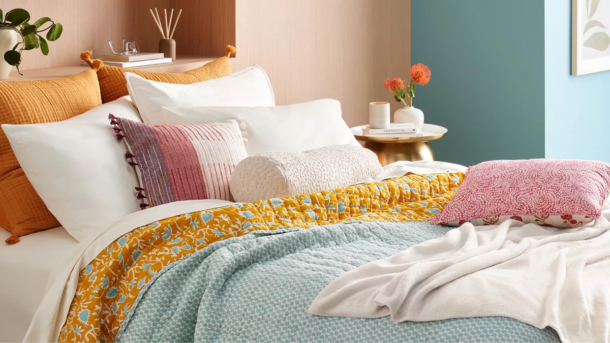 A light & airy bedroom has a layered bed with many textures & spring-y colors. Sun shines in from the right, where a side table has flowers, a book & a candle. A diffuser & plant sit on the built-in headboard nook, along with reading glasses & books.