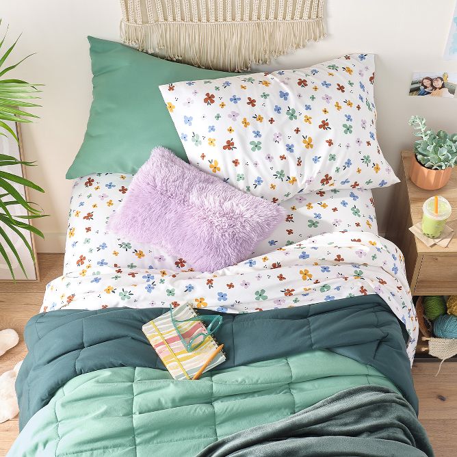 A sage green comforter is partially pulled down revealing floral print sheets. A notebook, glasses & pen are tossed on top of the bed with a fuzzy purple throw pillow. Above, a large macrame art piece hangs & plants sit on both sides of the room. 