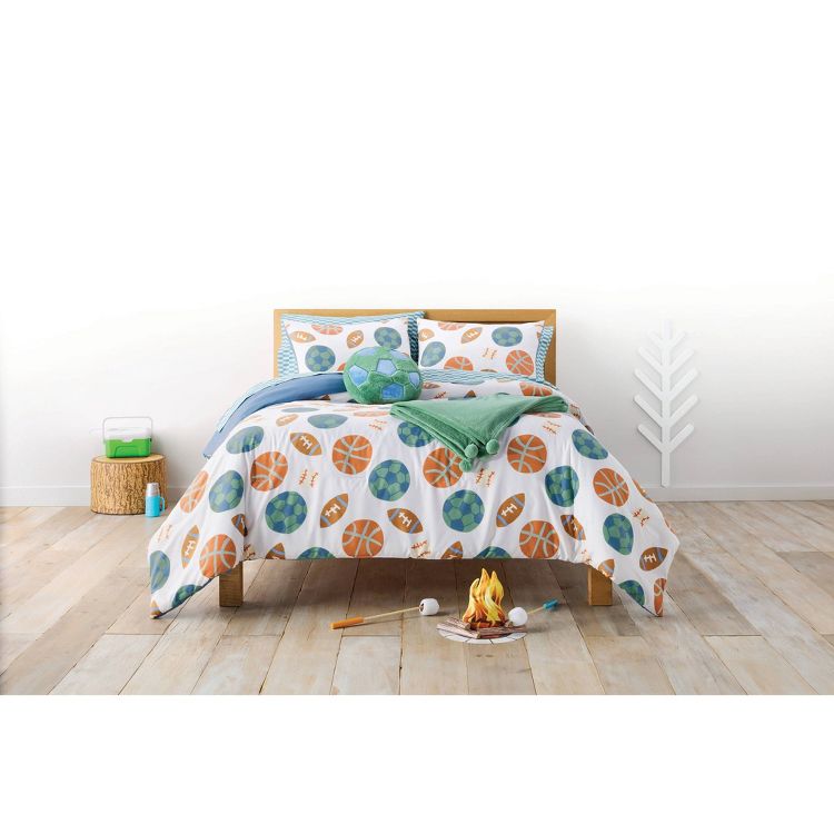 A bed with sports-themed bedding featuring basketball, soccer, and football designs, accompanied by a green towel and a plush soccer ball.