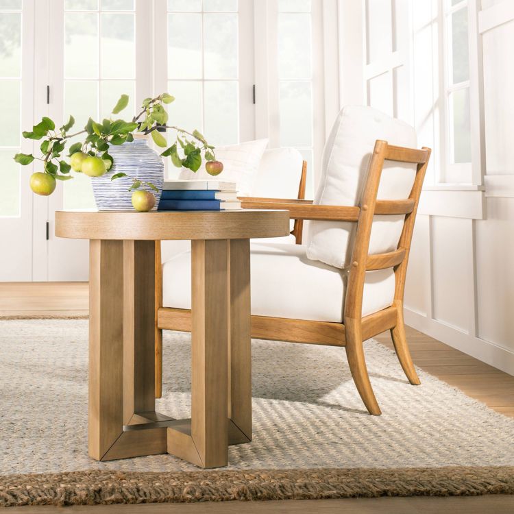 A round wooden table with a vase of apples and a stack of books on top, placed on a textured rug next to a cushioned wooden chair.