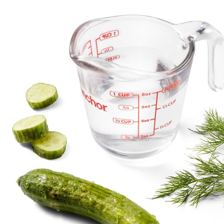 A clear measuring cup filled with liquid, marked with red measurements, next to sliced cucumber and dill.