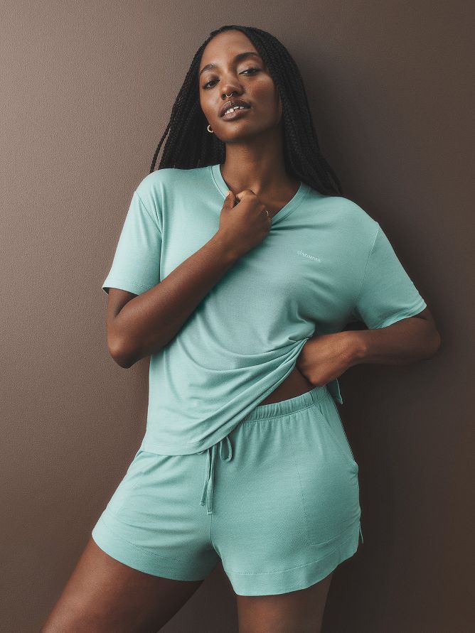 A woman is wearing an ultra-soft pajama set of aqua shorts with a tie and a matching aqua short-sleeve tee. The fabric is designed to help you relax and have a restful sleep. 