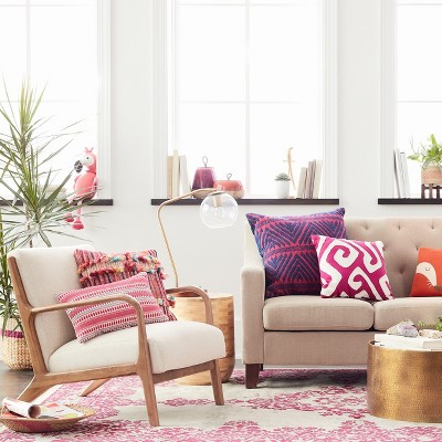 A beige colored armchair with wooden frame and a pink patterned cushion is placed on a pink patterned rug.
