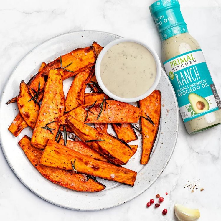 A bottle of Primal Kitchen Ranch dressing with avocado oil is placed next to a plate of roasted sweet potato wedges and a bowl of dipping sauce.